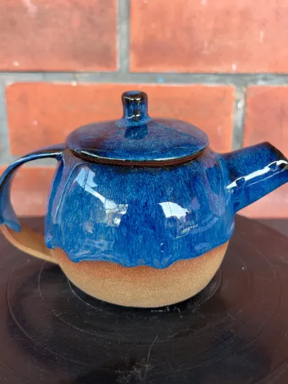 A wheel-thrown teapot by a student, featuring a deep blue dripping glaze over the natural stoneware clay body.