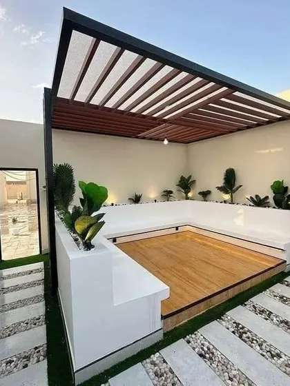 A clean and contemporary patio design with a white pergola, built-in seating, and a wooden deck. The space is surrounded by a pebble and stone pathway.