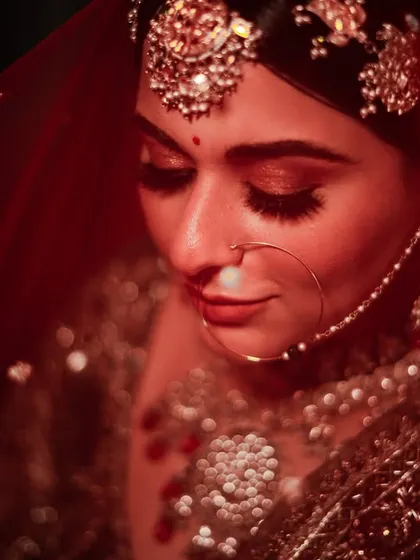 A close-up bridal portrait under a red veil. The lighting highlights her beautiful makeup and traditional jewelry, creating a dramatic and intimate feel.