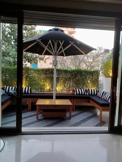 A view from the living room into a modern balcony seating area. The space is defined by an L shaped wooden bench, a large black umbrella, and a hedge of green plants for privacy.
