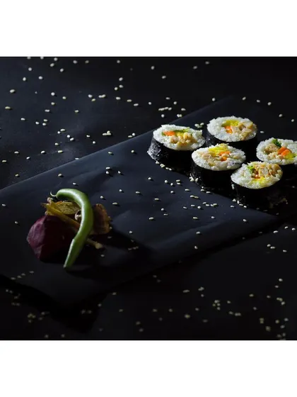 Minimalist plating of sushi on a black slate, with scattered sesame seeds creating a sense of movement. The focused lighting and dark background make this a refined shot for a Japanese restaurant menu.