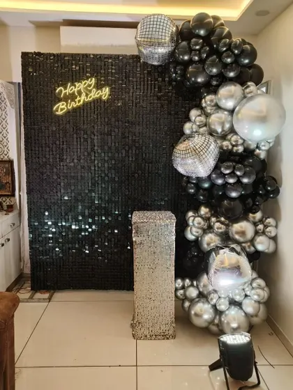 A chic and modern party setup with a black sequin wall. The decor includes a neon sign, a silver plinth, and a balloon arch made of black, silver, and disco ball balloons.