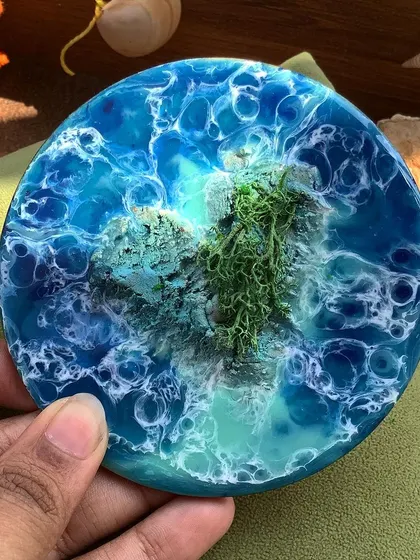 A handmade resin coaster with a green mossy island, held in my hand to show its size and detail.
