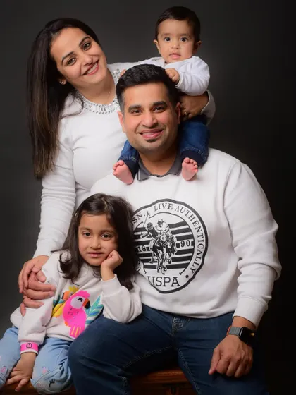 A beautiful family portrait showcasing coordinated outfits against a classic studio backdrop. The baby sitting on dad's shoulders adds a lovely, playful touch.