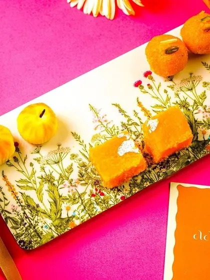 A rectangular serving platter styled with Indian sweets, shot on a vibrant pink background. The composition is clean and eye-catching.