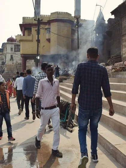 People walk along the steps of the Banaras ghats, with smoke from funeral pyres in the background. This image shows the continuous cycle of life and ritual by the Ganges.