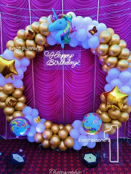 A classic unicorn-themed birthday decoration. This setup uses a gold and white balloon ring, accented with a unicorn foil balloon, star-shaped balloons, and a 'Happy Birthday' neon sign, perfect for a magical celebration.