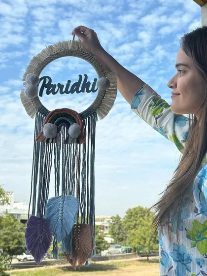 This custom macrame dreamcatcher for 'Paridhi' looks stunning against the sky. The design includes a nameplate, a mini rainbow, and multi-colored macrame feathers, making it a unique piece of wall art.