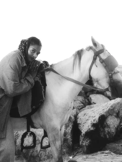 A powerful black and white fashion shot of a model leaning against a white horse amidst a rocky landscape. The high-contrast lighting and strong composition create a bold, editorial feel.