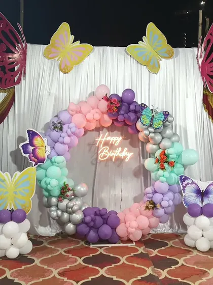 An outdoor butterfly-themed setup with a pastel balloon ring, neon sign, and large butterfly cutouts on balloon pillars.