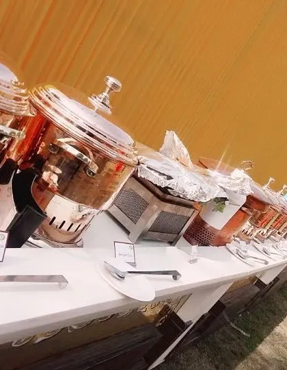 A classic outdoor buffet line set against a vibrant yellow marquee for a daytime event. The setup features a long table with our traditional copper and silver serveware, ready for guests.