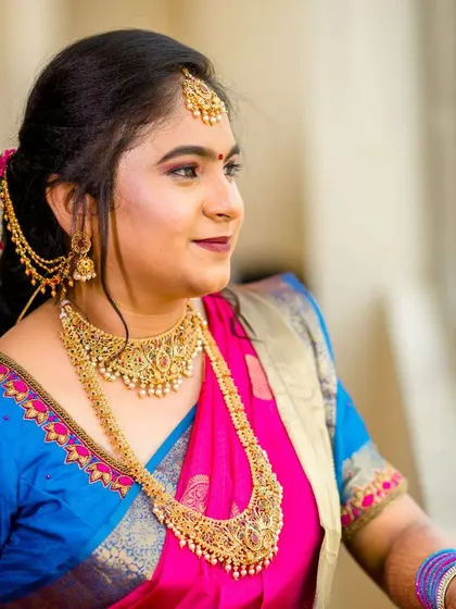 A candid moment of the bride, looking radiant. The makeup is set to be picture-perfect, with a focus on a smooth base and colors that complement her bright pink and blue saree.