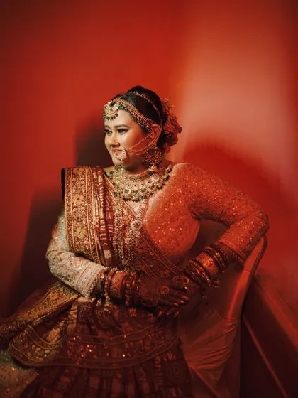A regal bridal portrait. The bride is seated, her posture exuding confidence and grace. The warm, dramatic lighting beautifully highlights her intricate jewelry and lehenga.