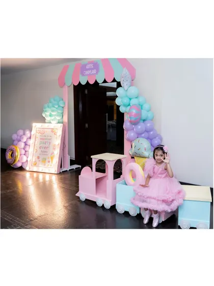 The birthday girl poses at the entrance to her sweet-themed party. The setup includes a candy-striped arch, a pink train, and a light-up welcome sign.