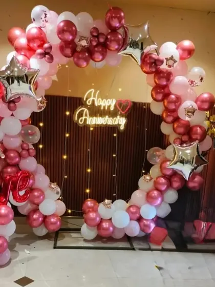 A beautiful anniversary decoration with a heart-shaped balloon frame. The setup uses red, pink, and white balloons, star-shaped foil balloons, and a 'Happy Anniversary' neon sign.
