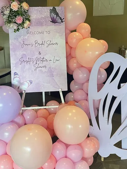 A welcome sign for a butterfly-themed bridal and mother-in-law shower. The sign is placed on an easel and surrounded by a pink and lavender balloon arrangement with a large butterfly cutout.