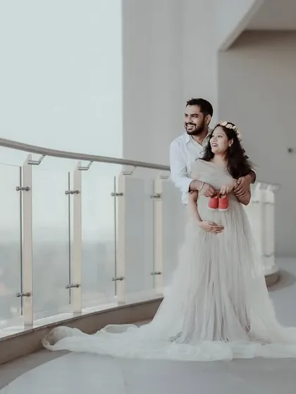 A wide shot of an expecting couple on a modern balcony, looking out at the view. The mother-to-be holds a pair of small baby shoes, symbolizing their future together as a family.