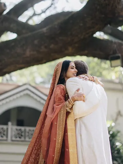 A warm embrace. The bride's rust-colored lehenga and the groom's ivory sherwani create a warm, earthy palette against the natural backdrop.