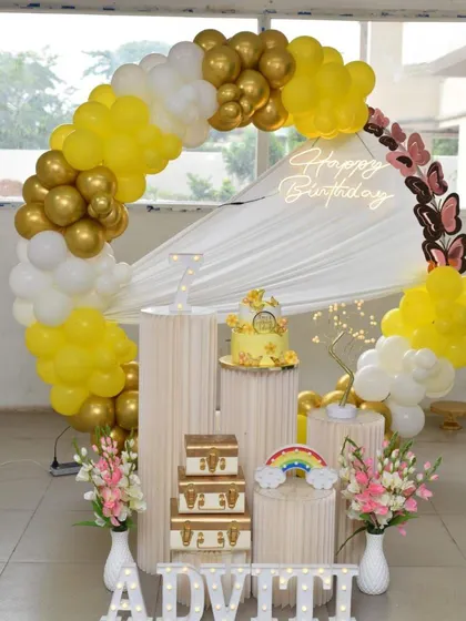 A bright and sunny birthday decoration with a yellow, white, and gold balloon arch. The setup includes decorative butterflies and a rainbow prop.