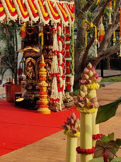 An outdoor wedding mandap setup that blends tradition with nature. The structure is heavily adorned with red, yellow, and white flower garlands, while lotus bud arrangements line the pathway.