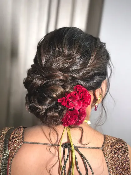This is a textured low bun adorned with deep red carnations. The messy yet controlled style provides a modern twist on the classic floral bun, suitable for a bride or bridesmaid.