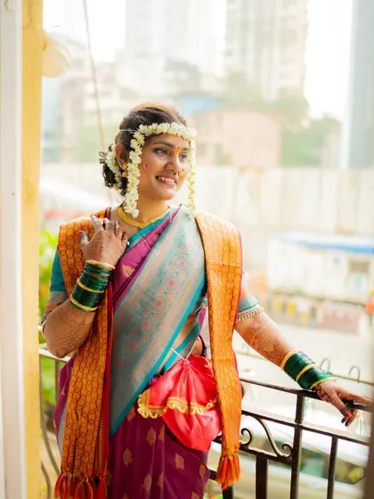 A beautiful portrait of the bride in a traditional Paithani saree, adorned with floral jewelry (gajra), looking out from a balcony.