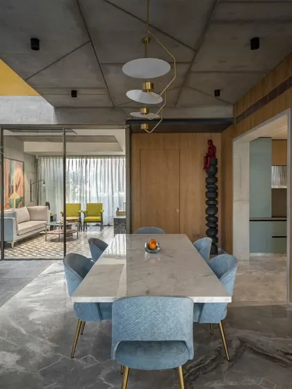 The dining area in Swatantra Residence serves as a focal point for visual connections between the courtyard, family sitting area, and kitchen. The blend of blue chairs, wooden walls, and a concrete ceiling creates an inviting and engaging environment.