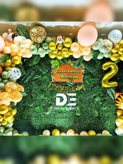 A full view of a jungle-themed birthday setup for Avyaan's second birthday. The decor features a lush green hedge wall, animal print balloons, and cutouts of a tiger and monkey.