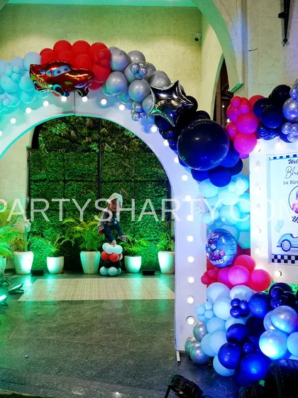 An illuminated entrance arch for a car party, decorated with balloons and a Lightning McQueen foil balloon, leading to a lush green backdrop.