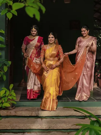 The bride, in a beautiful golden-yellow saree, is helped by her sisters as she walks.