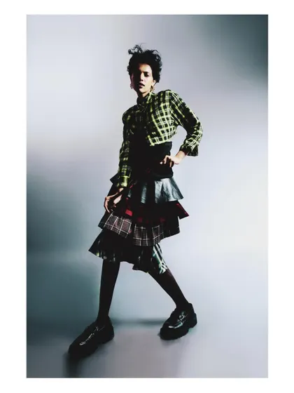 A full-length studio portrait from 'My Flatmate's Wardrobe,' featuring an eclectic, layered plaid outfit. The clean background and dynamic pose highlight the unique personal style celebrated in this series.