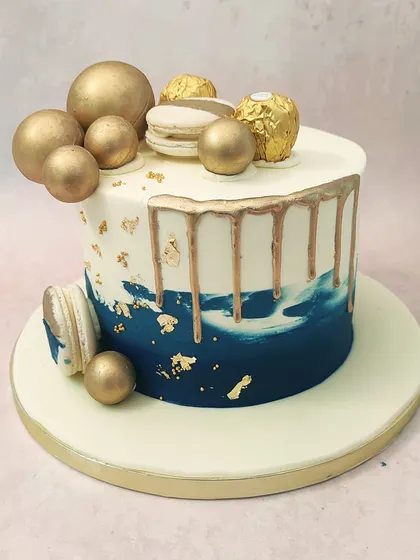 We design a cake of pure modern luxury with this blue and gold drip cake. A deep blue ombre effect is accented with gold drips, gold leaf, and an arrangement of golden spheres, Ferrero Rocher, and macarons, creating a sophisticated look for any celebration.
