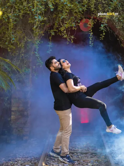 A playful and energetic shot of a couple on railway tracks, with vibrant blue smoke creating a dramatic and fun atmosphere.