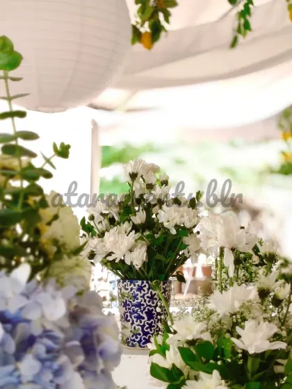 A close-up of a floral centerpiece at a Sicilian-themed party. The arrangement of white daisies and blue hydrangeas in a blue and white patterned vase perfectly complements the Mediterranean decor.