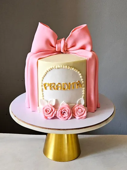 A classic and beautiful bow cake. This design features a perfectly draped pink fondant bow over a white ganache cake, accented with a pearl border and delicate sugar roses.