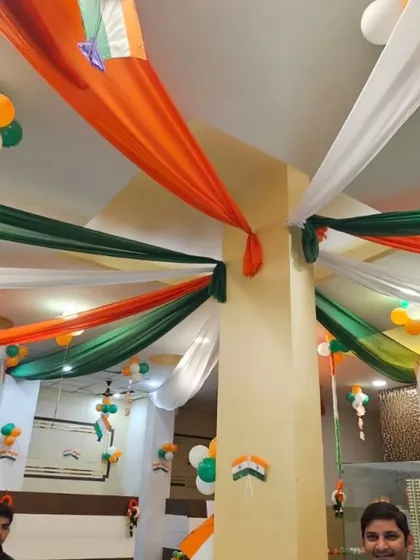 Ceiling decoration for an office cafeteria using saffron, white, and green fabric drapes and balloon clusters. This creates a festive and cheerful atmosphere for employees to celebrate together.