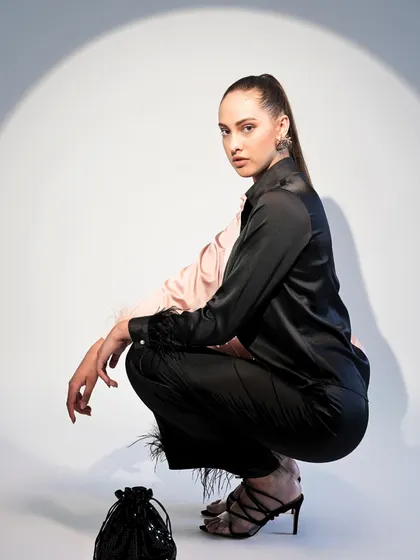 This look features a unique two-toned satin shirt with feather-trimmed cuffs. The crouching pose highlights the texture and detail of the trousers and shirt.