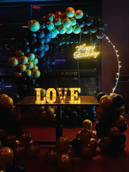 We can adapt our decorations to suit the mood of your venue. In this dimly lit space, a black and gold balloon arch with a 'Happy Birthday' neon sign and 'LOVE' marquee letters creates an intimate and celebratory atmosphere.