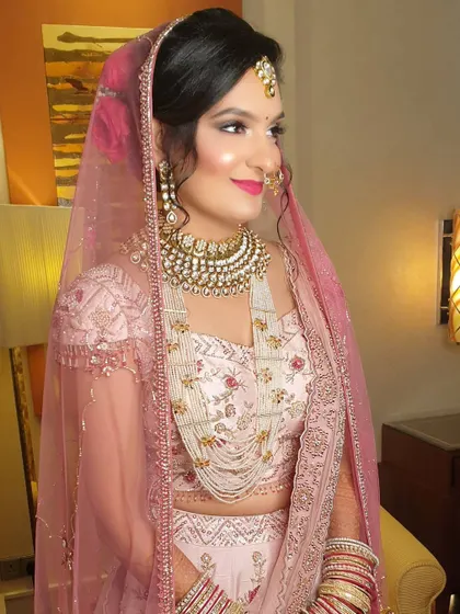 A profile shot of a bride in a delicate pastel pink lehenga. Her makeup is soft and romantic, with a pink lip and glowing skin, complemented by a heavy kundan necklace and a simple maang tikka.