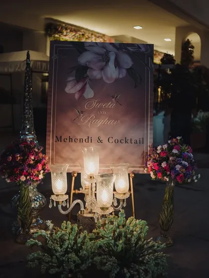 An elegant welcome sign for a 'Mehendi & Cocktail' night. The sign is surrounded by candles and floral arrangements, creating a romantic and sophisticated entrance.