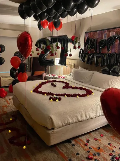 A bold and romantic 27th birthday hotel room decoration. This design uses black and red balloons, some with hanging roses, a heart made of petals on the bed, and large foil number balloons to mark the occasion.