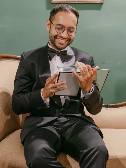 The groom, dressed in a tuxedo, smiles as he looks at a photo book, a thoughtful gift or part of a speech during the reception.