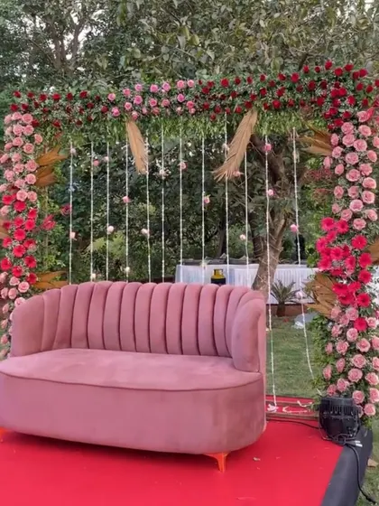 A romantic outdoor seating area for a pre-wedding event. A pink sofa is set against a backdrop of a floral arch and hanging lights, creating a perfect photo spot.