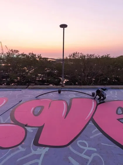Working on a large pink "LOVE" piece on the roof of the skatepark at sunset. This shows the process of creating large-scale lettering with rollers and spray cans.