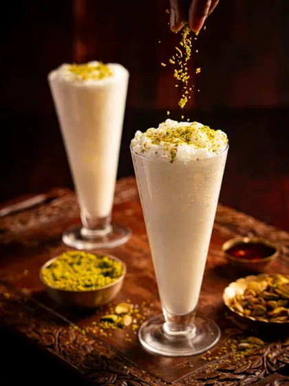 A tall glass of lassi or falooda from Gallops, with pistachios being sprinkled from above. The action shot adds a final, delicious touch to this traditional drink.