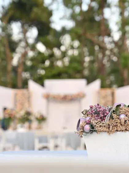 The beauty of pastel centerpieces. This close-up shows a delicate arrangement of dried lavender and other muted florals, with the elegant outdoor wedding stage blurred softly in the background.