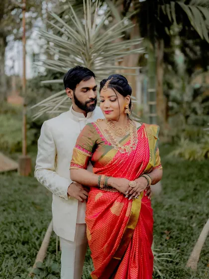 A classic pose that showcases both the couple's bond and their beautiful wedding attire. The lush green background provides a perfect contrast to the bride's vibrant red saree.