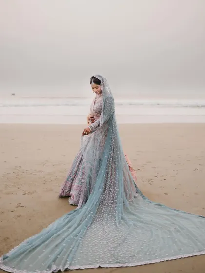 A bride stands on a serene beach at dusk, her magnificent light blue veil trailing behind her, creating a breathtaking and ethereal bridal portrait against the ocean.