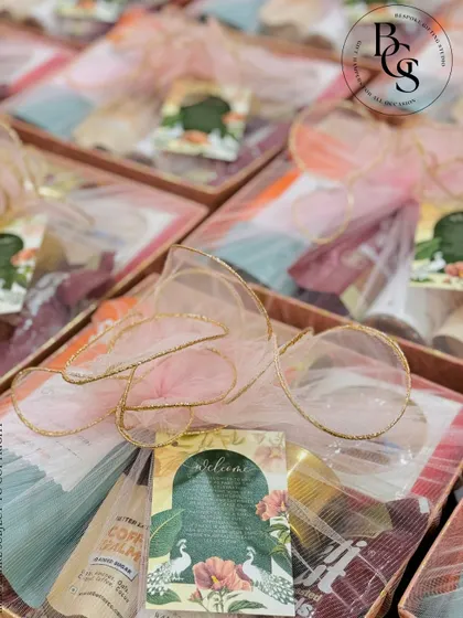 A welcome hamper for a wedding, featuring delicate pastel fabric wrapping. The sheer organza is tied with a gold-trimmed ribbon, and a custom welcome card with a peacock motif is tucked in, setting a beautiful tone for the event.