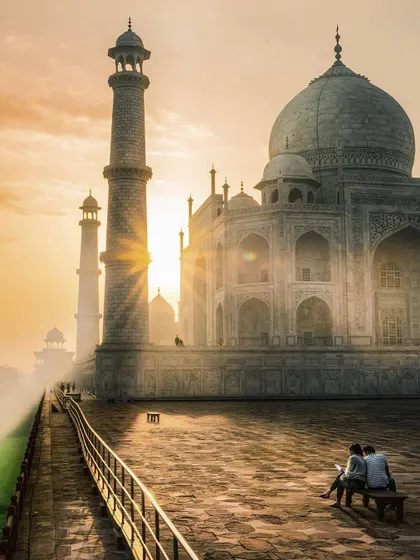 The morning sun casts a warm, hazy glow over the Taj Mahal, with a couple sitting quietly in the foreground. This is a classic and romantic view of one of the world's most famous monuments.